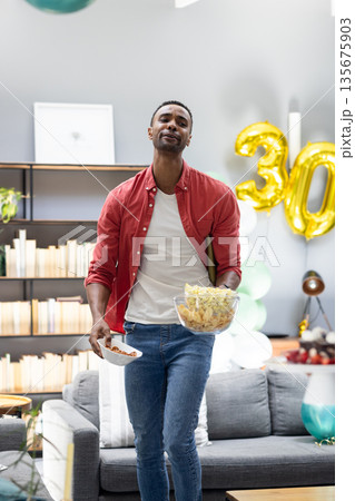 Man in red shirt carrying snacks at home party with balloons in background 135675903