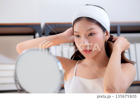 Young asian woman wearing headband and tying hair while preparing skincare looking mirror. Young asian woman wearing headband and tying hair while preparing skincare looking mirror. 135675920