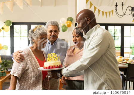 Elderly woman blowing out candles on birthday cake, surrounded by joyful friends 135676014
