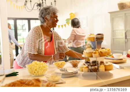 Elderly woman arranging cupcakes on table, smiling during family celebration at home 135676169