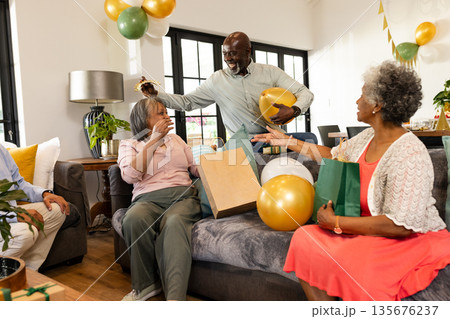 Elderly friends celebrating birthday at home with gifts and balloons, sharing joy 135676237