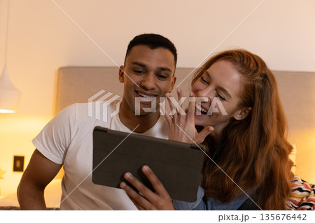 Young couple laughing while watching tablet together in cozy bedroom Young couple laughing while watching tablet together in cozy bedroom 135676442