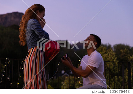 Young man proposing with ring to surprised woman on romantic evening balcony Young man proposing with ring to surprised woman on romantic evening balcony 135676448