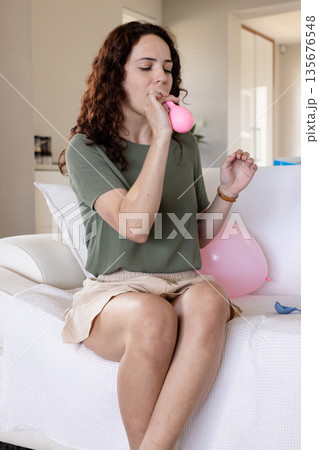 Inflating pink balloon, woman sitting on couch at home, preparing for celebration Inflating pink balloon, woman sitting on couch at home, preparing for celebration 135676548