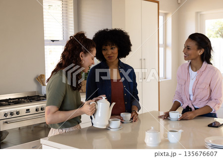 Women enjoying conversation while pouring tea in modern kitchen, smiling warmly 135676607
