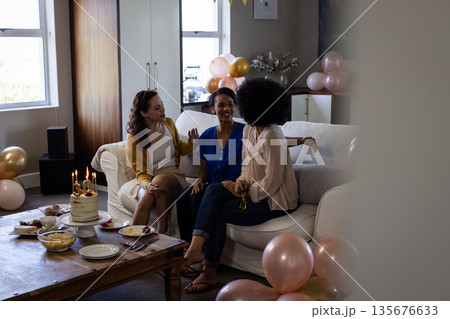 Women sitting on sofa celebrating birthday with cake and balloons at home 135676633