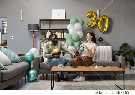 Couple celebrating birthday at home, laughing and holding balloons on sofa Couple celebrating birthday at home, laughing and holding balloons on sofa 135676656