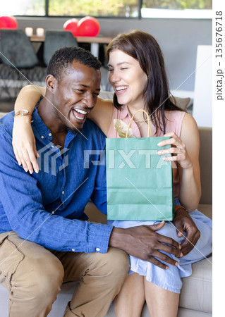 Couple celebrating with gift bag, laughing together on cozy living room sofa Couple celebrating with gift bag, laughing together on cozy living room sofa 135676718