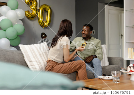 Celebrating 30th birthday, couple having cake and chatting on cozy living room sofa Celebrating 30th birthday, couple having cake and chatting on cozy living room sofa 135676791