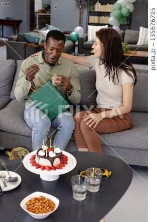 African American man opening gift with woman smiling beside him at home celebration African American man opening gift with woman smiling beside him at home celebration 135676795