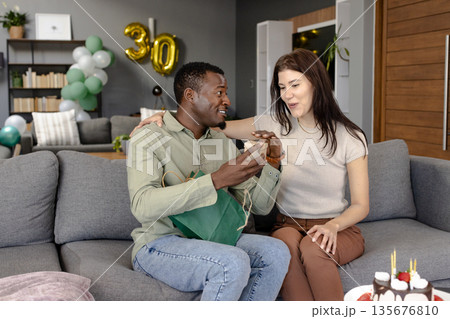 Celebrating birthday, African American man giving gift to woman on living room sofa 135676810