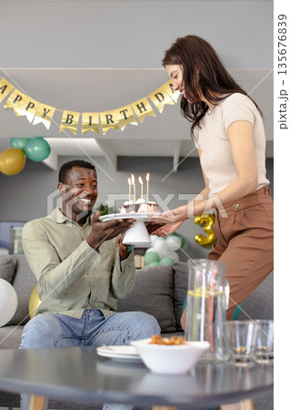Smiling man receiving birthday cake from woman at home celebration with balloons Smiling man receiving birthday cake from woman at home celebration with balloons 135676839