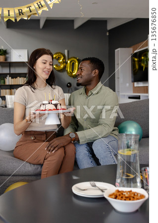 Celebrating birthday, woman presenting cake to smiling African American man on couch Celebrating birthday, woman presenting cake to smiling African American man on couch 135676850