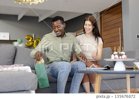 African American man opening gift bag on couch, celebrating birthday with friend African American man opening gift bag on couch, celebrating birthday with friend 135676872