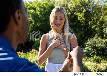 Man proposing to surprised woman in garden, holding ring box with joy Man proposing to surprised woman in garden, holding ring box with joy 135676877