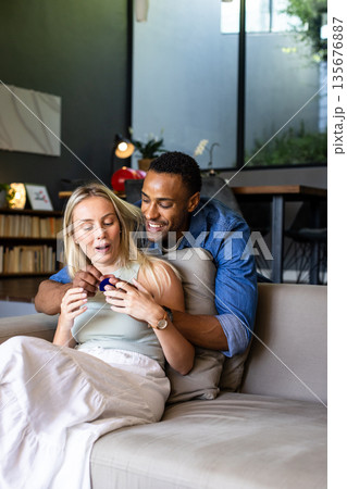 Couple at home, man surprising woman with gift, both smiling and joyful 135676887