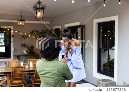 Man proposing to surprised woman on patio, romantic moment under string lights 135677228