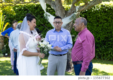 Holding bouquet, bride in white dress smiling with guests outdoors 135677302