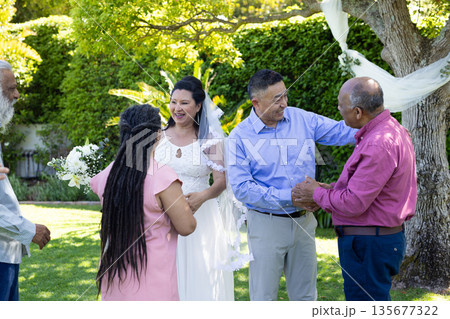 Congratulating newlywed couple, seniors under decorated tree during outdoor celebration 135677322