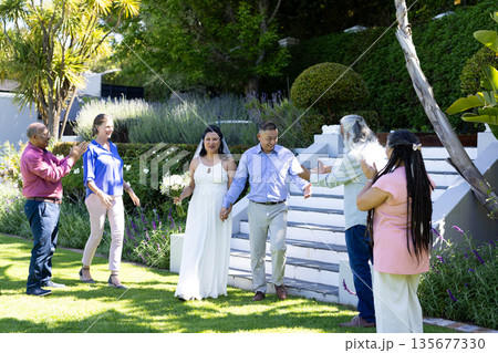 Walking hand in hand, bride and groom surrounded by joyful family outdoors Walking hand in hand, bride and groom surrounded by joyful family outdoors 135677330