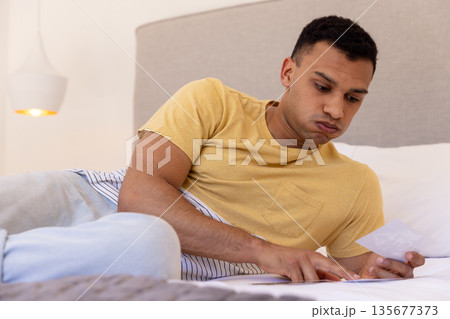 Young man lying on bed looking at photograph, appearing thoughtful and relaxed Young man lying on bed looking at photograph, appearing thoughtful and relaxed 135677373