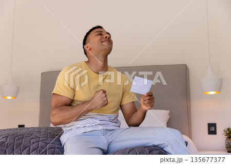 Young man sitting on bed celebrating while holding photograph, feeling joyful Young man sitting on bed celebrating while holding photograph, feeling joyful 135677377