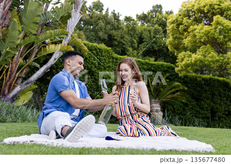 Young couple enjoying picnic outdoors, sharing champagne and smiling on blanket Young couple enjoying picnic outdoors, sharing champagne and smiling on blanket 135677480