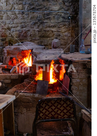 : A traditional glass furnace with bright flames and glowing embers. The structure is made of stone and metal, showcasing the glass-making process. 135678704