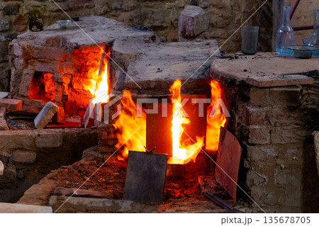 : A traditional glass furnace with bright flames and glowing embers. The structure is made of stone and metal, showcasing the glass-making process. 135678705