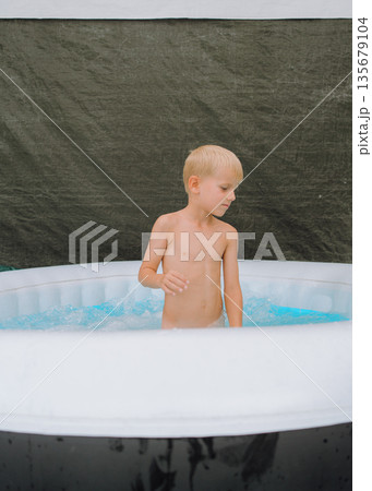 Young boy stands in an inflatable pool filled with water. The child appears to be enjoying the moment, with a thoughtful but relaxed expression, engaging with the surrounding water. 135679104