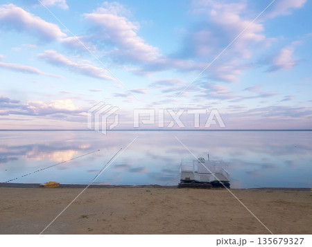 Calm water of lake reflecting sunset sky with clouds. Serene coastal landscape for travel and tranquility. Peaceful nature scene. Calm water of lake reflecting sunset sky with clouds. Serene coastal landscape for travel and tranquility. Peaceful nature scene. 135679327