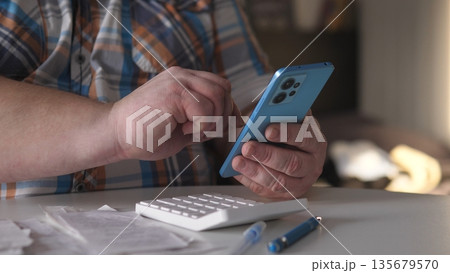 A person is focused on a smartphone while seated at a table. Nearby, a calculator and various papers are visible. The setting has soft light coming from outside. A person is focused on a smartphone while seated at a table. Nearby, a calculator and various papers are visible. The setting has soft light coming from outside. 135679570