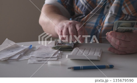 A person sorts cash while using a calculator on a table covered with receipts and a pen. The setting shows a focus on managing money in a quiet space during day time. A person sorts cash while using a calculator on a table covered with receipts and a pen. The setting shows a focus on managing money in a quiet space during day time. 135679590