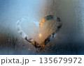 A heart-shaped clear patch melted into frosty glass, glowing warmly against cold blue condensation. 135679972