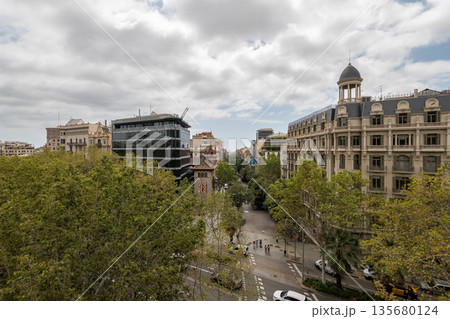 Urban landscape of Barcelona Eixample district featuring a mix of modern and historical architecture. Green trees line the street under a cloudy sky. Cars are parked along the road. 135680124