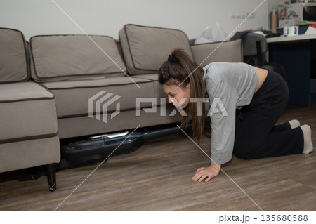 Woman on Hands and Knees Uses Vacuum to Clean Under the Sofa at Home 135680588