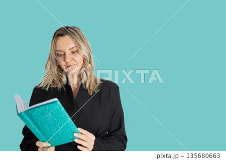 Woman in Business Suit Attentively Reads a Turquoise Book on a Plain Background 135680663