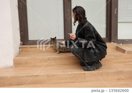 Woman kneels on stairs to gently greet a curious cat outside a cozy home setting during a calm afternoon Woman kneels on stairs to gently greet a curious cat outside a cozy home setting during a calm afternoon 135682600