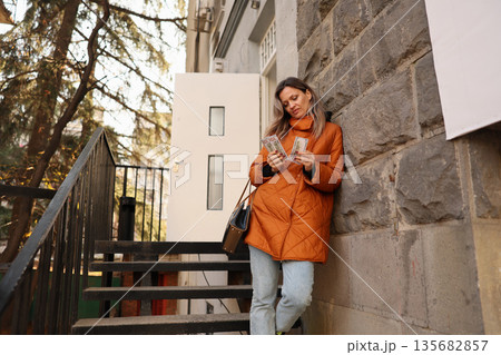 Busy afternoon in the city as a young woman enjoys her time while checking her phone near a stone building Busy afternoon in the city as a young woman enjoys her time while checking her phone near a stone building 135682857