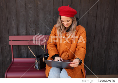 Young woman in a red beret enjoys her time on a laptop while sitting on a bench outside a cozy wooden building Young woman in a red beret enjoys her time on a laptop while sitting on a bench outside a cozy wooden building 135682910