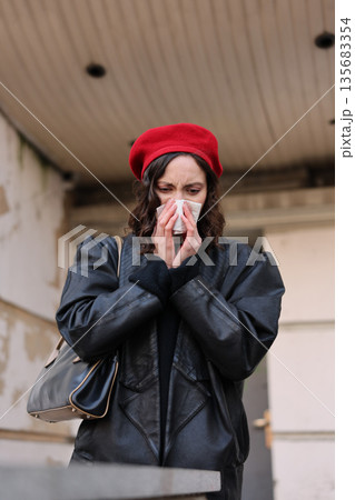 Young woman wearing a red beret and black coat sneezes while standing outside a building in autumn Young woman wearing a red beret and black coat sneezes while standing outside a building in autumn 135683354