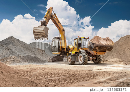 A loader transports sand from the ground to a storage bin at a construction site. An excavator assists. Heavy equipment works with the soil on a clear day 135683371