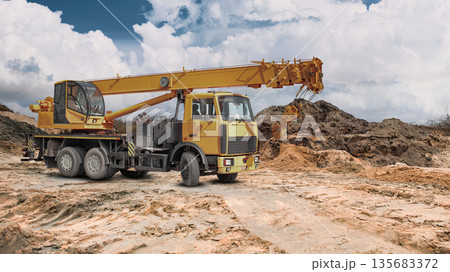 A truck crane stands ready for work at a construction site in a vast area filled with earth embankments, under a partially cloudy sky 135683372