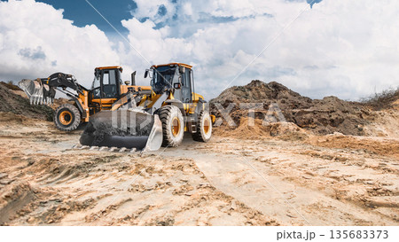 Two powerful wheel loaders stand ready to work in a sand quarry, while clouds disperse across the blue sky. The equipment is ready for construction work 135683373