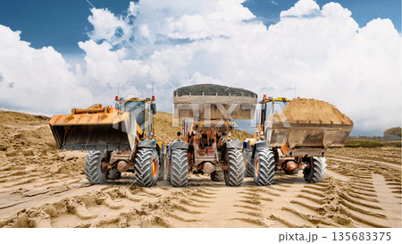 Three powerful loaders with raised buckets stand at the construction site, workers continuing construction on a cloudy day. Large piles of sand indicate the ongoing work 135683375