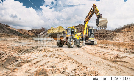 A construction loader moves soil, while in the background an excavator with its bucket raised. Clouds are visible in the sky, creating a contrast with the daytime landscape 135683388