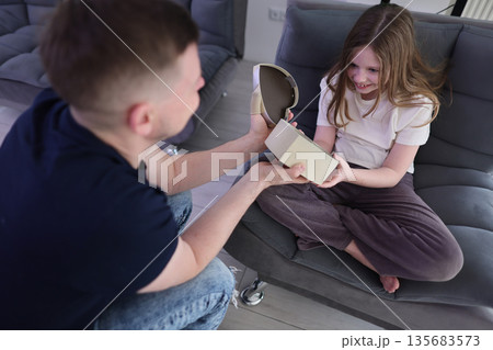 Joyful child opening a surprise gift at home with her smiling dad during a cozy afternoon Joyful child opening a surprise gift at home with her smiling dad during a cozy afternoon 135683573