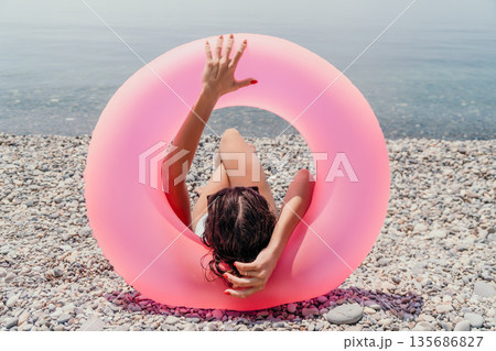 Beach summer vacation woman rests behind a large pink inflatable ring while lying on a rocky shore by the water 135686827