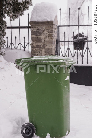 Green Recycling Bin Covered in Snow Near Stone Pillar and Black Iron Gate in Winter Landscape Green Recycling Bin Covered in Snow Near Stone Pillar and Black Iron Gate in Winter Landscape 135687811