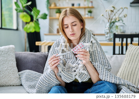 Sad upset teenage girl looking at thermometer holding blister pack of pills sitting at home Sad upset teenage girl looking at thermometer holding blister pack of pills sitting at home 135688179